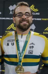 Man with glasses smiling holding a gold medal for cycling
